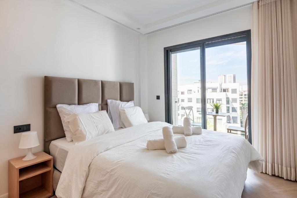 A hotel room with a large bed, a nightstand, and a view of the city through a large window.