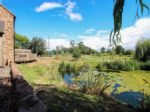 Ein Steinhaus mit einer Holzterrasse, einem Teich mit Seerosen und einem eingezäunten Bereich mit Bäumen und Gras.