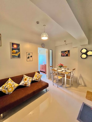 A modern living room with a brown sofa, dining table, and chairs. There are wall-mounted artworks and a ceiling light fixture.