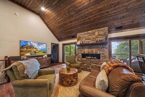 A spacious living room with a stone fireplace, comfortable seating, and a large window offering a view of the outdoors. - Luxury Cabin with Hot Tub & Mountain Views (Zionville)