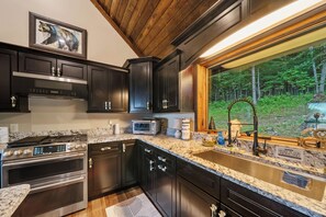 A modern kitchen with dark cabinetry, stainless steel appliances, and a large window offering a view of a forest. - Luxury Cabin with Hot Tub & Mountain Views (Zionville)