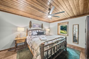 Ein Schlafzimmer mit Holzbalkendecke, einem Bett mit Tagesdecke im Bergmotiv, zwei Nachttischlampen, einem Fenster mit Blick ins Grüne und einem Spiegel an der Wand.