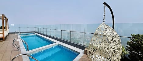 A rooftop pool with clear blue water, a glass balustrade, and a hanging wicker chair.