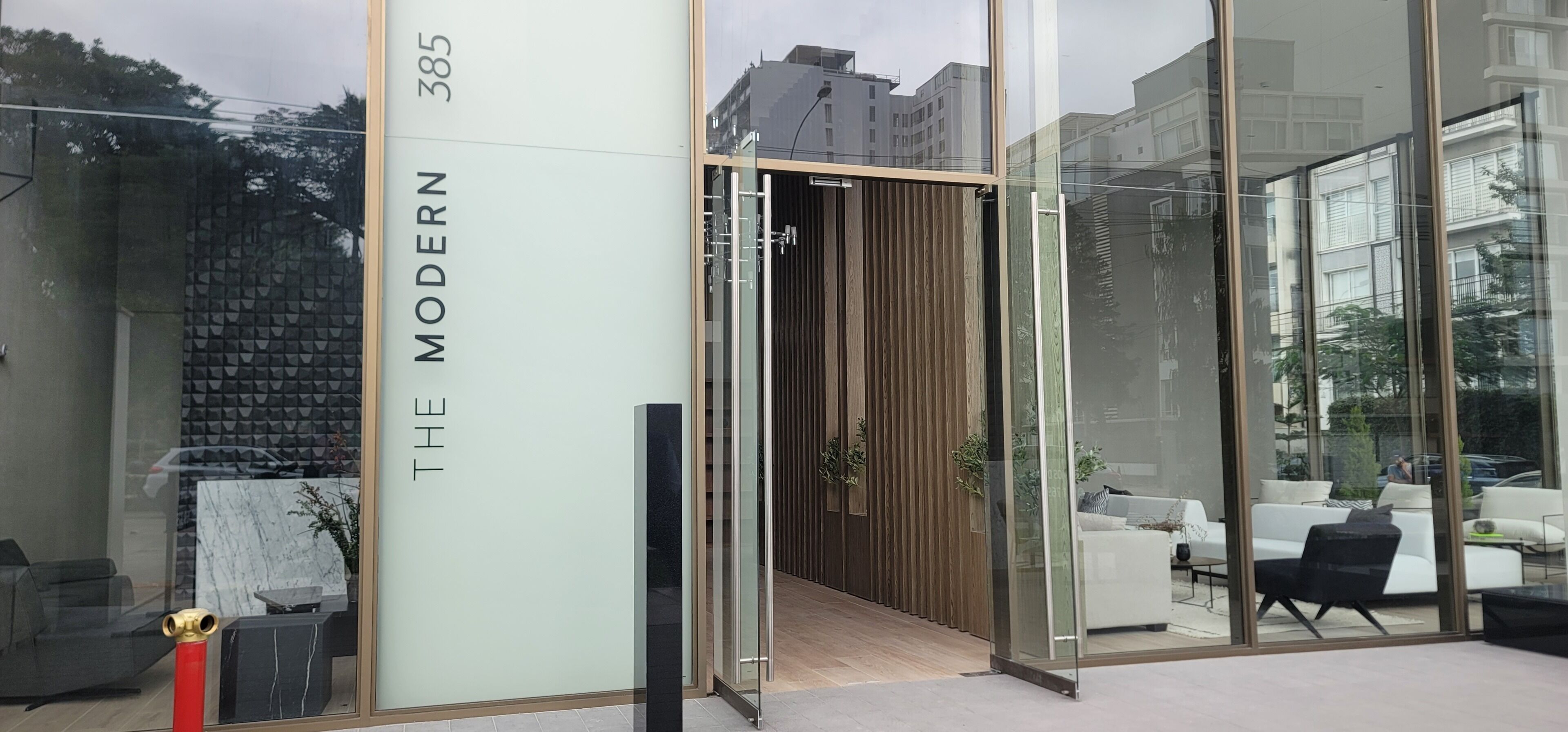 A modern building entrance with a glass facade, a wooden interior, and a visible outdoor seating area.