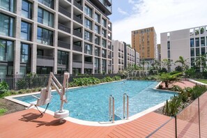 A swimming pool with a wooden deck, surrounded by a fence, and adjacent to a multi-story building with balconies.