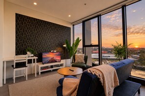 A modern living room with a large TV, a sofa, a coffee table, and a view of the city at sunset.