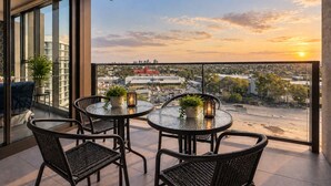 En balkon med gelænder af glas, møbleret med fletstole og runde borde, med udsigt over et bybillede i solnedgang.