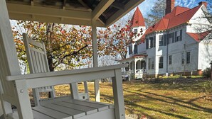 A white rocking chair on a porch with a view of a white house with a red roof.