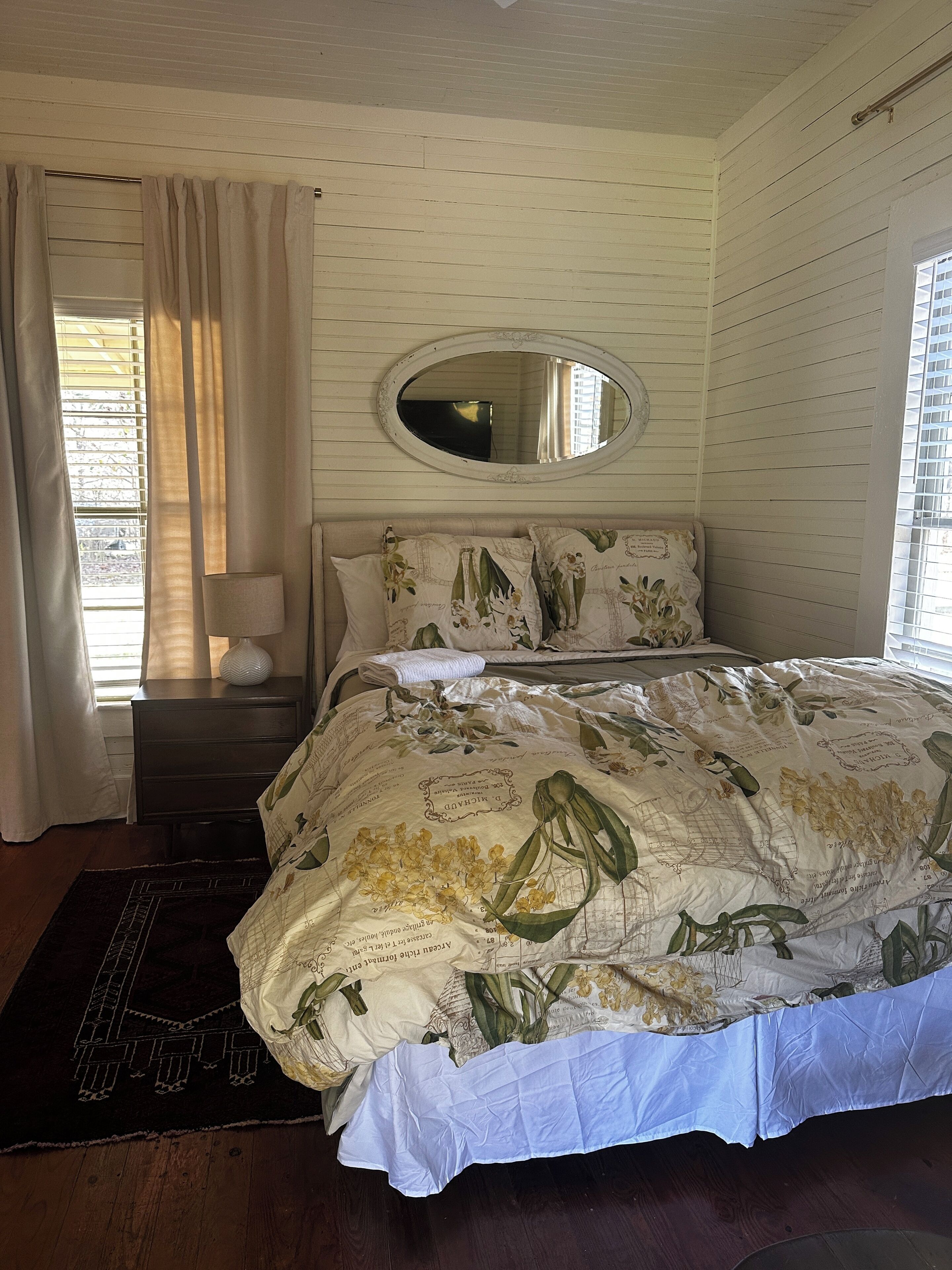 A bedroom with a bed, a nightstand, a mirror, and a window with curtains.