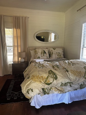A bedroom with a bed, a nightstand, a mirror, and a window with curtains. - Historic tiny cottage in Waynesville peaceful location near downtown (Waynesville)