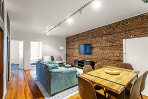 A modern living space with a brick wall, a sofa, a dining table with chairs, and a flat-screen TV mounted on the wall. - Hells Kitchen 3BR, Walk to Times Square, Broadway & The Vessel (New York)