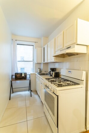 A compact kitchen with white cabinets, a gas stove, a microwave, and a coffee maker. - Hells Kitchen 3BR, Walk to Times Square, Broadway & The Vessel (New York)