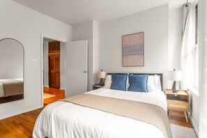 A hotel room with a bed, bedside tables, a mirror, a wardrobe, and a painting on the wall. - Hells Kitchen 3BR, Walk to Times Square, Broadway & The Vessel (New York)