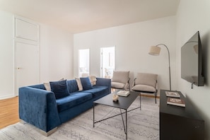 Ein modernes Wohnzimmer mit einem blauen Sofa, einem Couchtisch und zwei Sesseln. An der Wand ist ein Flachbildfernseher angebracht.