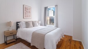 Ein Schlafzimmer mit Bett, Nachttischen, einem Fenster mit Vorhängen und einem gerahmten Bild an der Wand.
