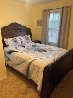 A bedroom with a bed, a nightstand, a window with curtains, and a painting on the wall.