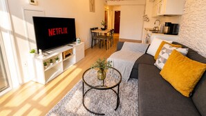 Ein Wohnzimmer mit einem Flachbildfernseher, der 'NETFLIX' anzeigt, eine Essecke mit Tisch und Stühlen, eine Küche mit Steinrückwand und ein graues Sofa mit gelbem Kissen.