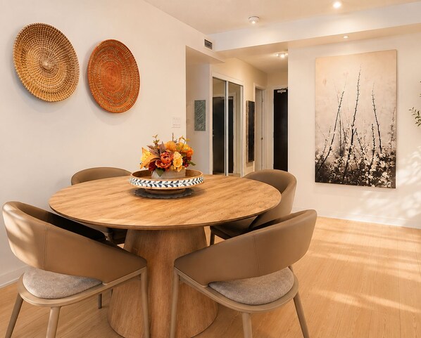 A dining area with a round wooden table, six chairs, a vase with flowers, two woven wall decorations, and a large framed picture on the wall.
