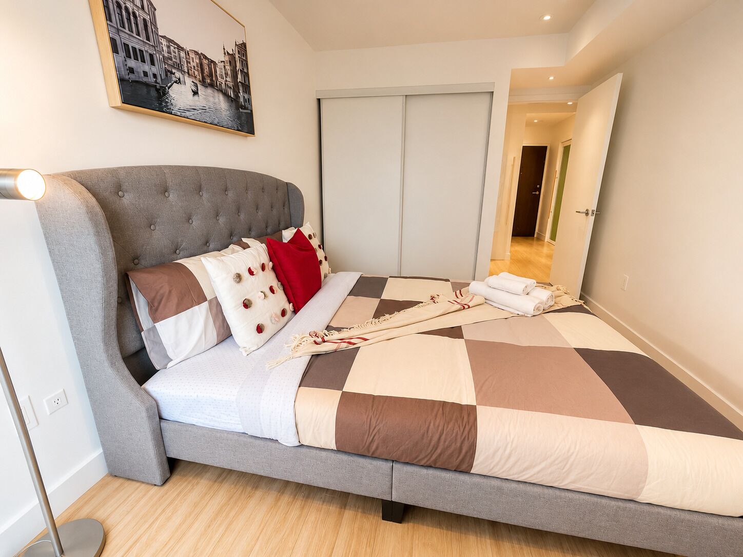A neatly made bed with a checkered bedspread, a grey upholstered headboard, and a wall-mounted picture of a cityscape.