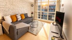 Ein modernes Wohnzimmer mit grauem Sofa, Couchtisch, Fernseher und einem großen Fenster mit Blick auf Gebäude.