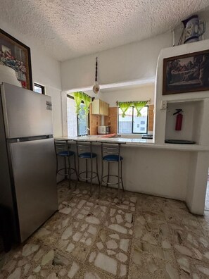 A kitchen with a refrigerator, bar stools, a microwave, and a fire extinguisher.