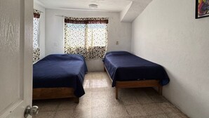 Une chambre avec deux lits, un ventilateur de plafond, une fenêtre avec des rideaux à motifs et un tableau au mur.