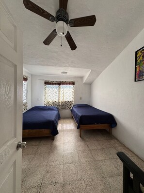 A room with two beds, a ceiling fan, a window with patterned curtains, and a painting on the wall.
