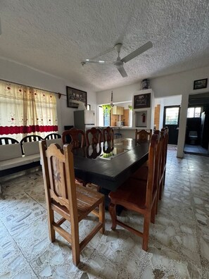 A dining area with a round table and wooden chairs, a ceiling fan, and a kitchen in the background.