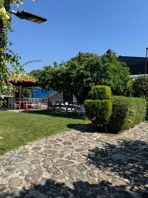 A paved outdoor area with a picnic table and chairs, surrounded by greenery and a clear blue sky.