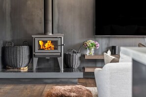 A modern living room with a wood-burning stove, wicker baskets, a vase with flowers, and a flat-screen TV mounted on the wall.