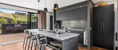 A modern kitchen with a central island, bar stools, and a view of a garden through large windows.