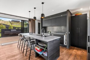 A modern kitchen with a central island, bar stools, and a view of a garden through large windows. - Luxury Hāwea | Alfresco Retreat | Sleeps 6 (Lake Hāwea)