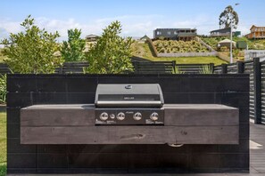 An outdoor grill with a control panel, set against a backdrop of a landscaped garden and residential buildings. - Luxury Hāwea | Alfresco Retreat | Sleeps 6 (Lake Hāwea)