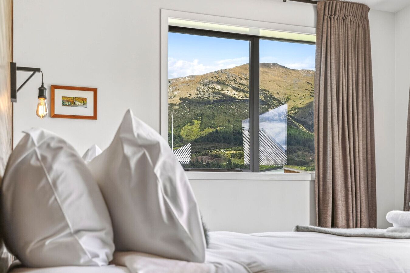 A hotel room with a bed, pillows, a framed picture, a window with a view of a mountain, and a curtain.