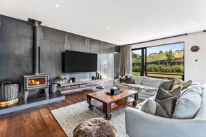 A modern living room with a fireplace, a flat-screen TV, a sofa, and a large window offering a view of the outdoors. - Luxury Hāwea | Alfresco Retreat | Sleeps 6 (Lake Hāwea)