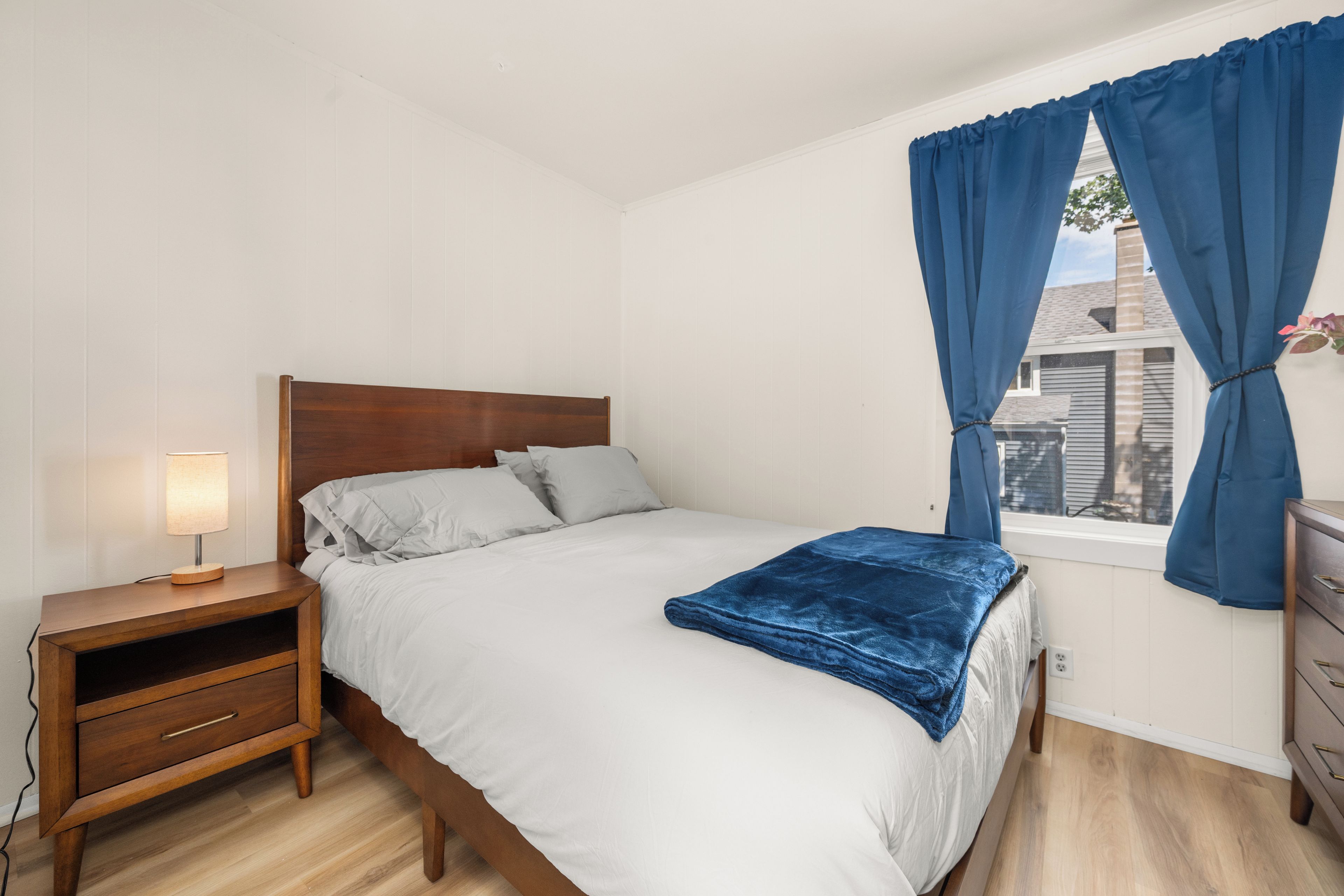 A bedroom with a wooden bed, white bedding, a bedside table with a lamp, and a window with blue curtains.