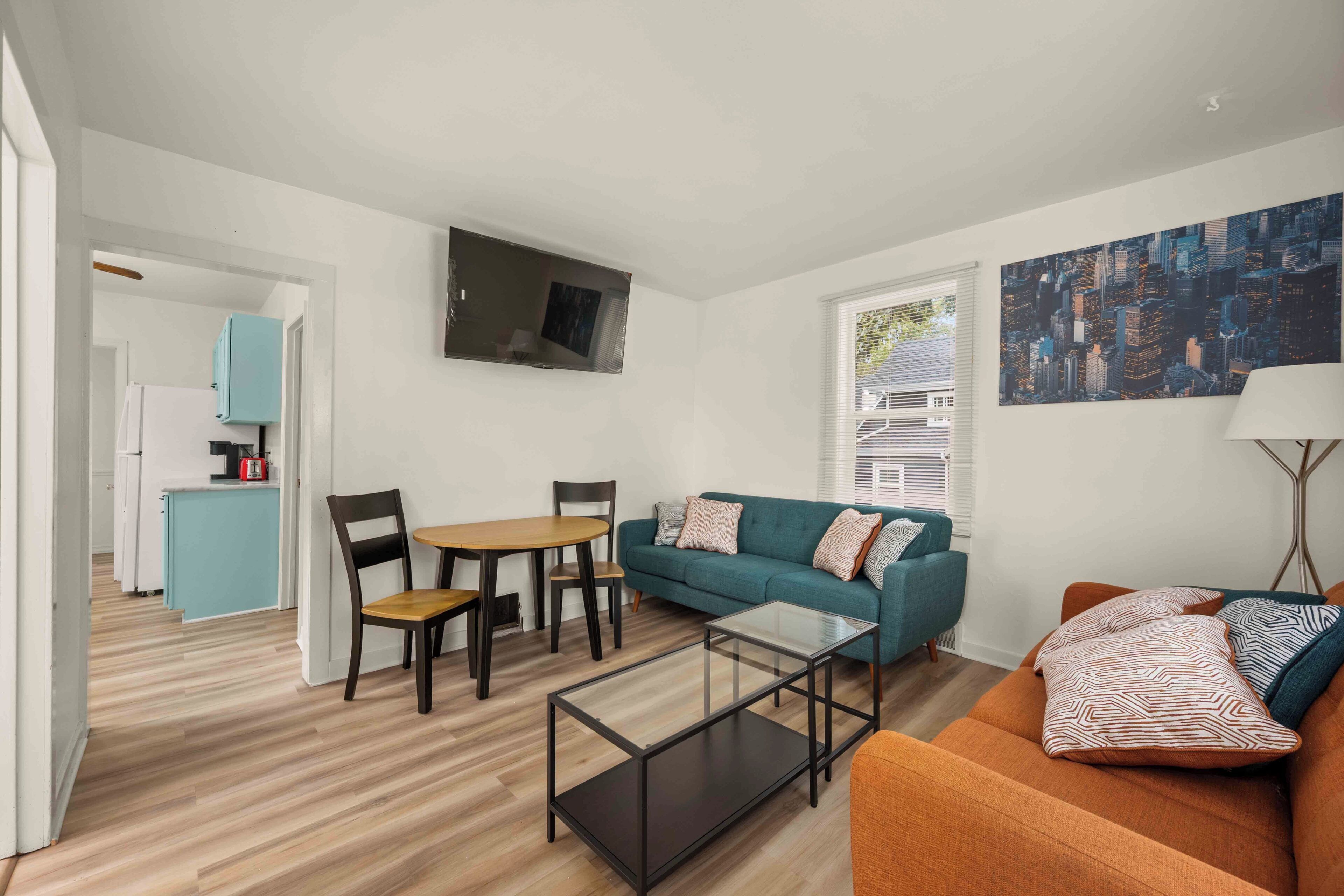 A modern living room with a sofa, coffee table, and chairs. There is a kitchen area with cabinets and a refrigerator. A flat-screen TV is mounted on the wall.