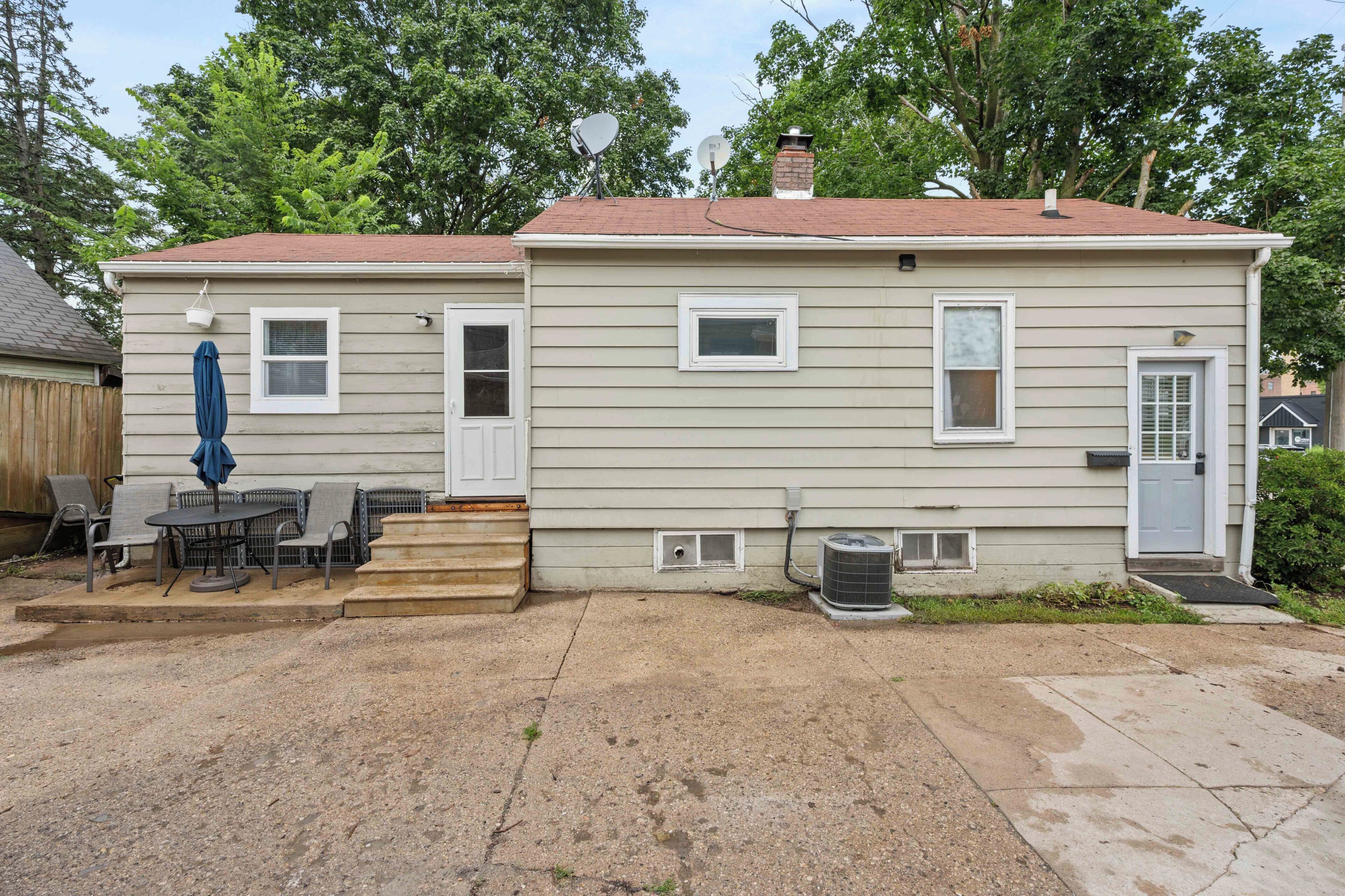 A single-story house with a front door, two windows, a satellite dish, and a small patio with outdoor furniture.