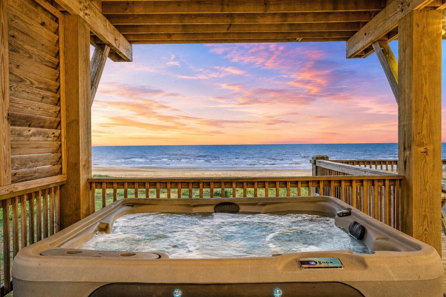 A wooden deck with a hot tub overlooking a beach at sunset.