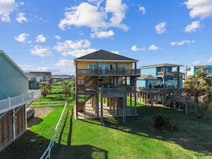 A multi-level wooden structure with balconies, surrounded by a grassy yard and palm trees, under a clear blue sky. - Beachfront Home in Crystal Beach | Private Hot Tub | Double Decks | Outdoor Bar | Sleeps 16 (Crystal Beach)