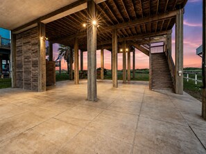 An open-air covered patio with a view of the sunset, wooden pillars, and a staircase leading to an upper level. - Beachfront Home in Crystal Beach | Private Hot Tub | Double Decks | Outdoor Bar | Sleeps 16 (Crystal Beach)