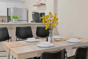 Un coin repas avec une table dressée pour quatre personnes, des chaises, un vase rempli de fleurs jaunes et, au fond, des appareils de cuisine.