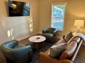 A living room with a flat-screen TV mounted on the wall, a brown leather sofa, two blue armchairs, a round coffee table, and a window with blinds. - Game Day Nest (Oxford)