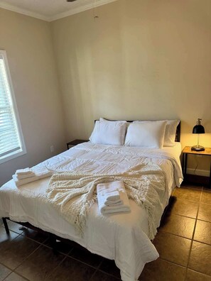 A neatly made bed with white linens and a folded towel, a bedside lamp, and a window with blinds. - Game Day Nest (Oxford)