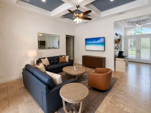Un salon avec un canapé bleu foncé, une table basse ronde et un fauteuil marron. On y trouve un ventilateur de plafond, un grand miroir et un tableau représentant une scène de plage.
