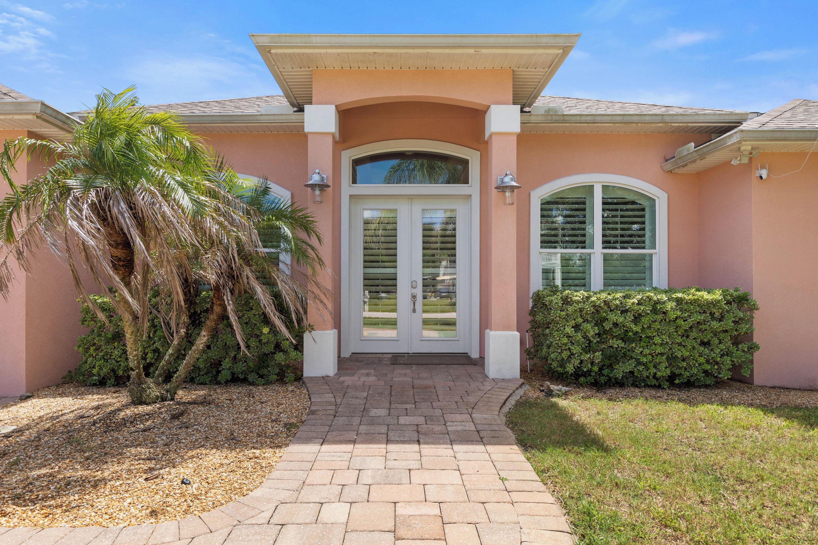 A residential house with a brick pathway leading to a double door entrance, flanked by a palm tree and a hedge.