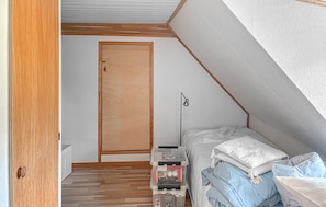 A small, narrow room with a sloped ceiling, a wooden door, a bedside table with a lamp, and a bed with pillows.