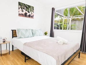 Un dormitorio con cama, almohadas, una mesita con una botella de agua, una planta y una ventana con cortinas.