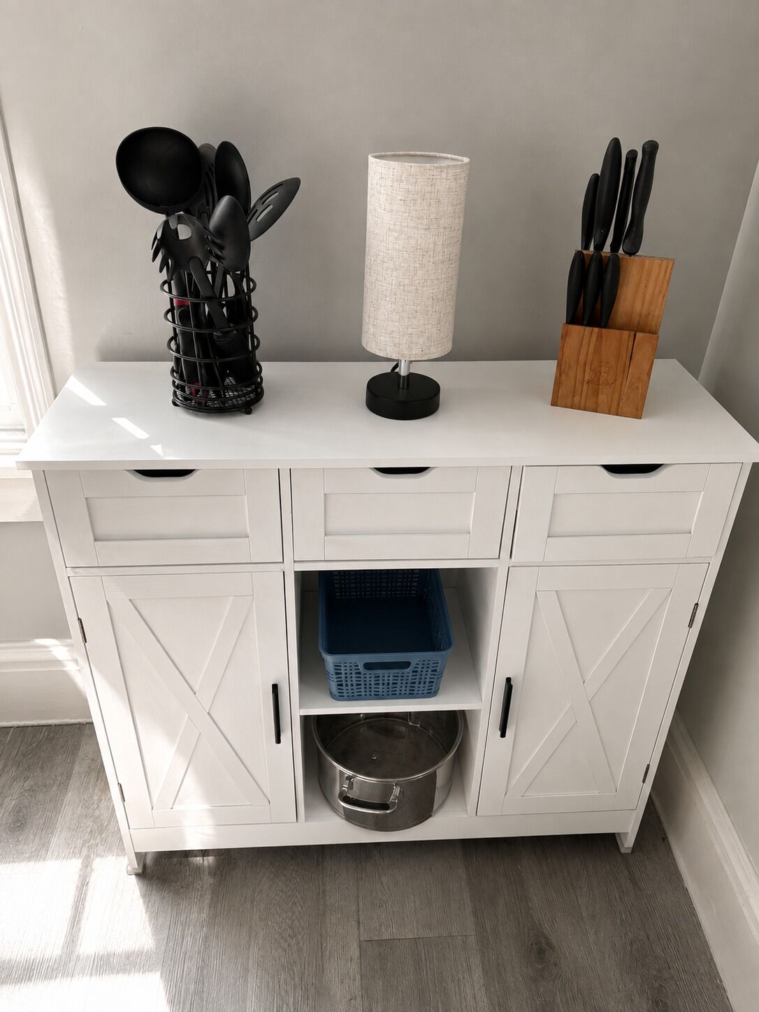 A white kitchen cabinet with a metal pot, a blue basket, and a black utensil holder.