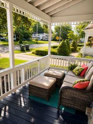 A covered porch with wicker furniture and cushions, a green mat, and a view of a suburban street with houses and a railroad crossing sign. - 3-bedroom house in lovely Painesville built in 1904! (Painesville)